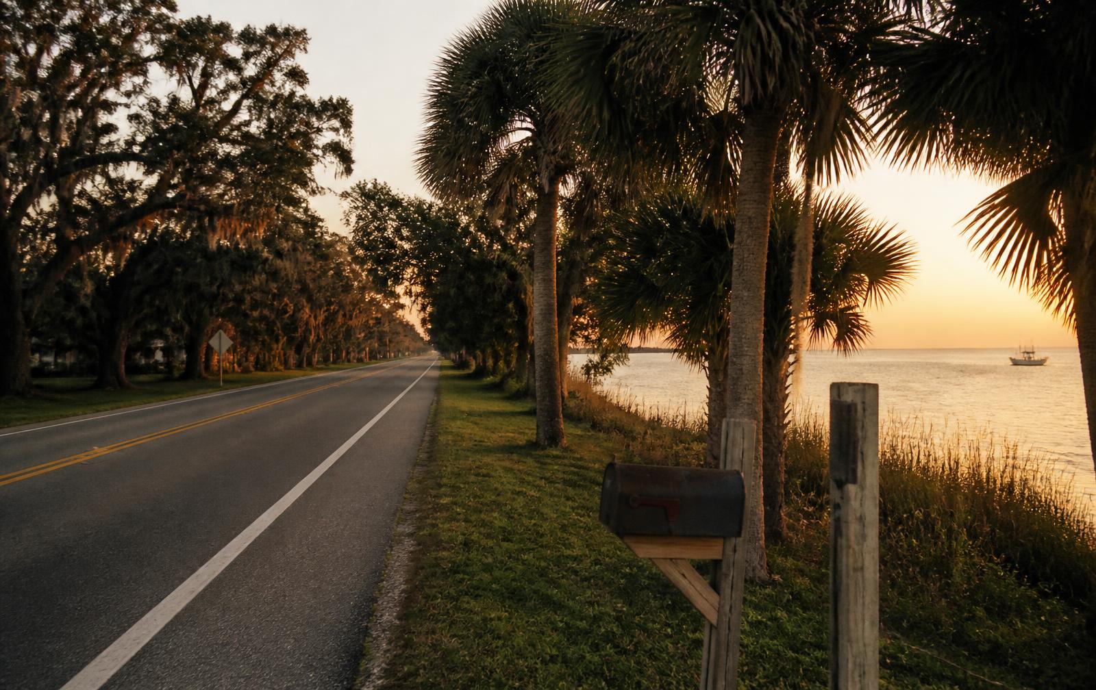 Empty two-lane road lined with cabbage palms and moss-draped oaks beside the Indian River Lagoon at golden hour near Grant-Valkaria, Florida